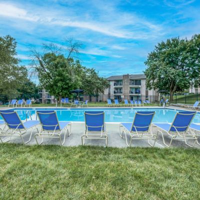 Outdoor swimming pool surrounded by blue lounge chairs, with trees and apartment buildings in the background.