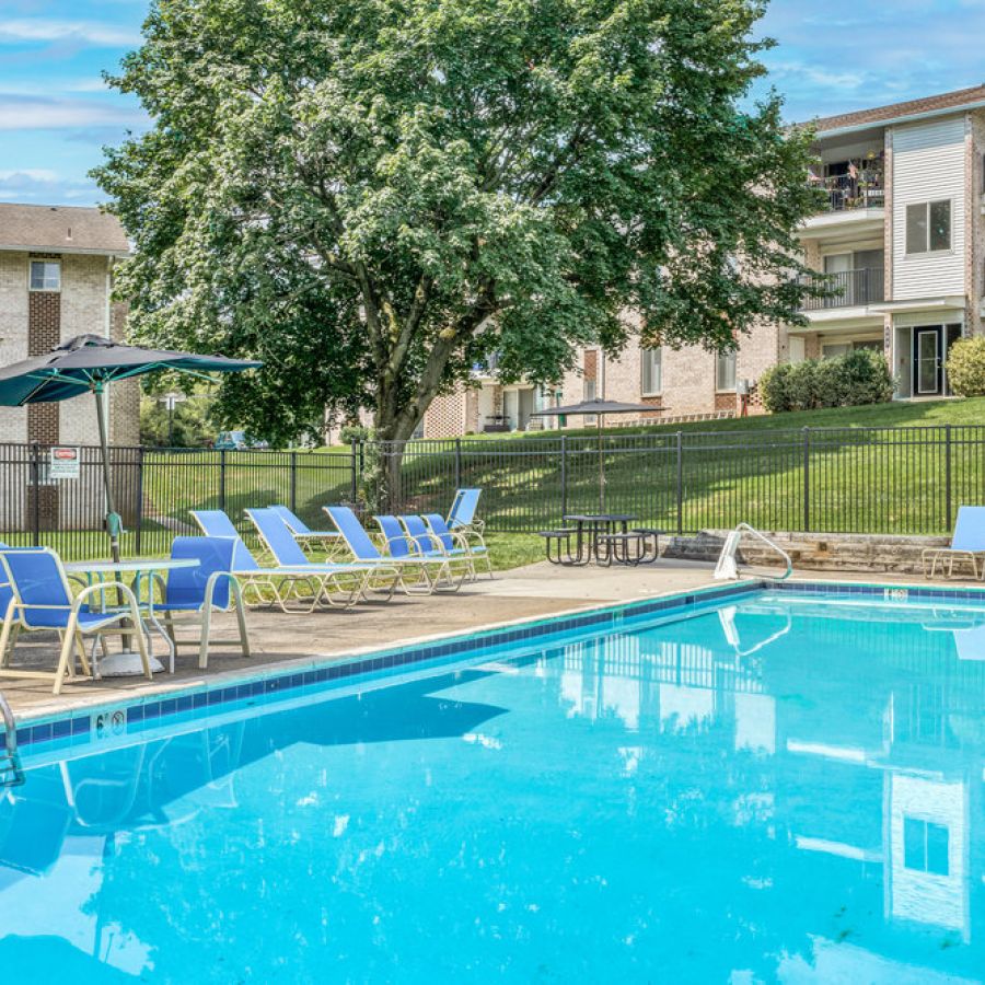 Outdoor swimming pool with blue lounge chairs, umbrellas, and apartment buildings in the background.