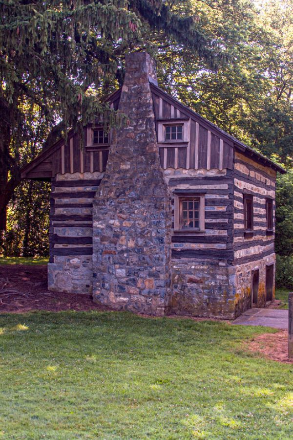 A small stone and wood log cabin sits beside a large tree in a grassy, wooded area on a sunny day.